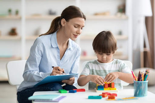 A child development specialist observes a child playing with blocks and takes notes on a clipboard.