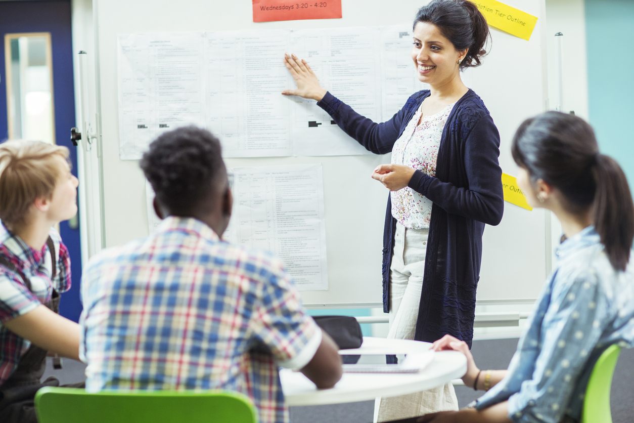 A teacher giving a lecture points to a whiteboard.