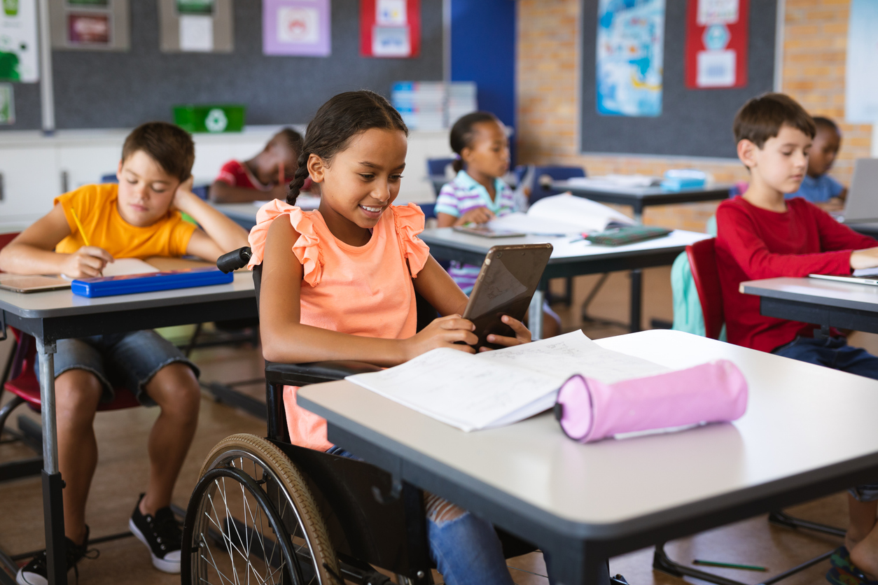 A Student in a Wheelchair Sits at a Desk at the Front of the Classroom and Works on a Tablet.
