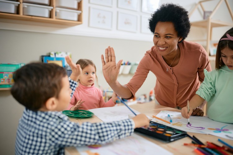 A smiling kindergarten teacher interacts with students creating artwork.