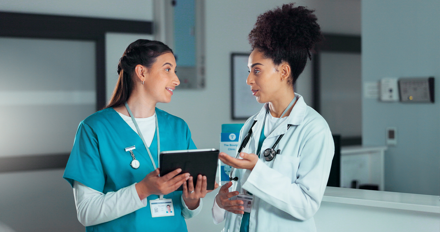 An APRN wearing a white coat talks to a nurse in scrubs.