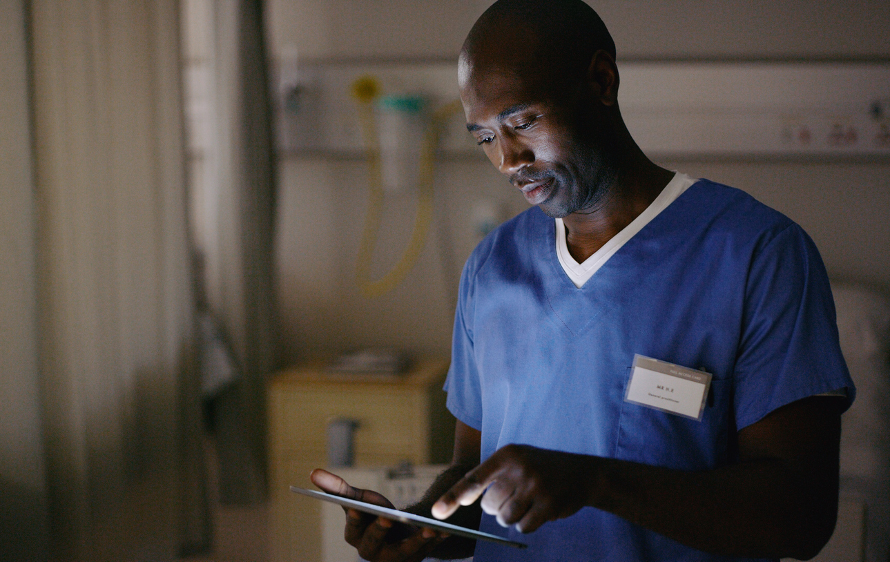 A nurse wearing scrubs in a hospital room making notes on a tablet. 