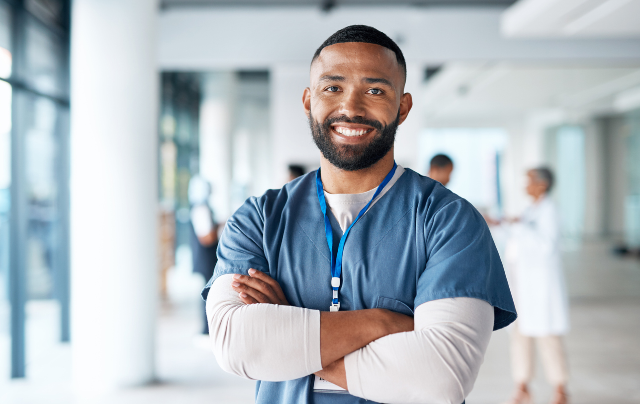 A smiling caregiver wearing scrubs standing in a hospital hallway.