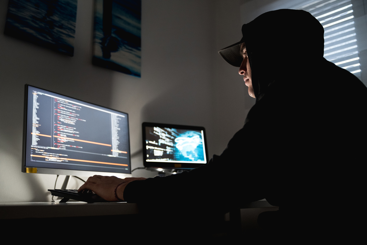 A criminal in a baseball cap and a hoodie works at a computer monitor in a darkened room.