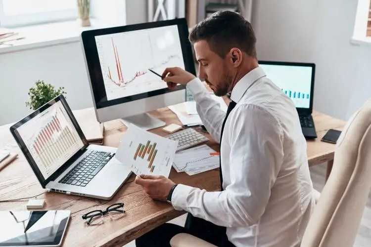 Businessperson Comparing Charts at a Desk With Three Computers