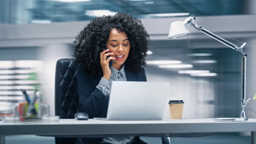 A professional sitting at a desk in an office talks on the telephone while looking at a laptop.