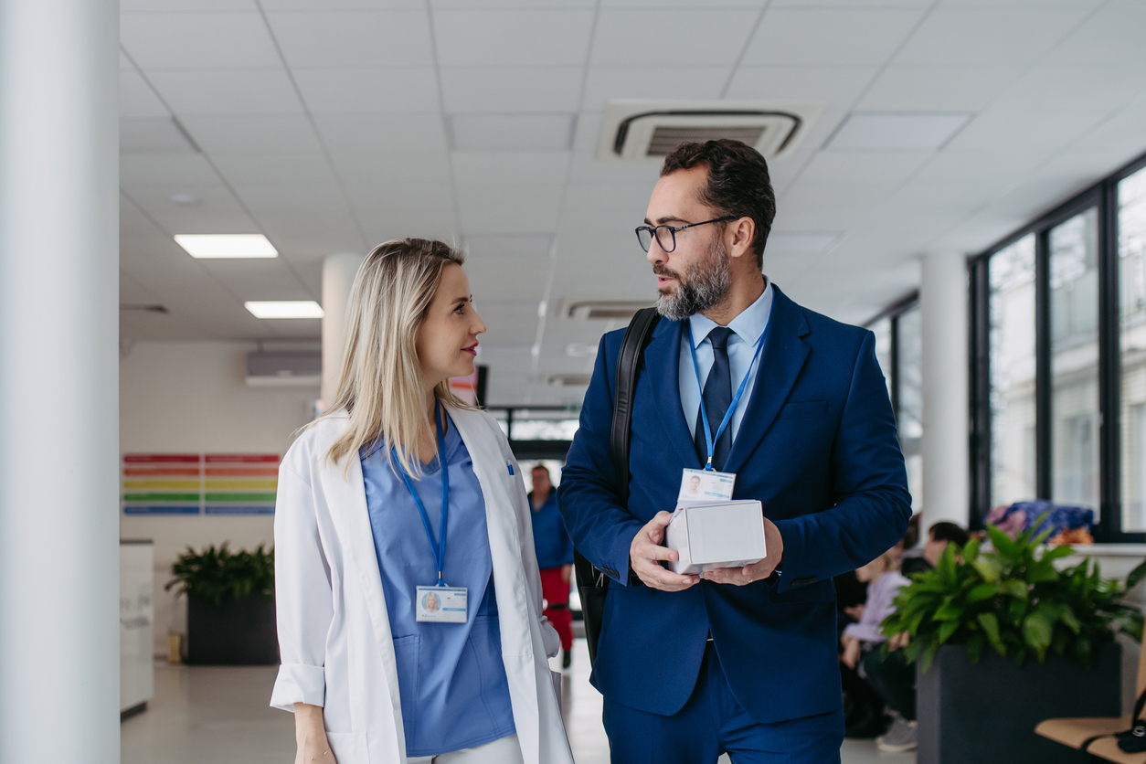 A Pharmaceutical Sales Rep Holding a Box of Samples Talks With a Physician in a Hospital Hallway.