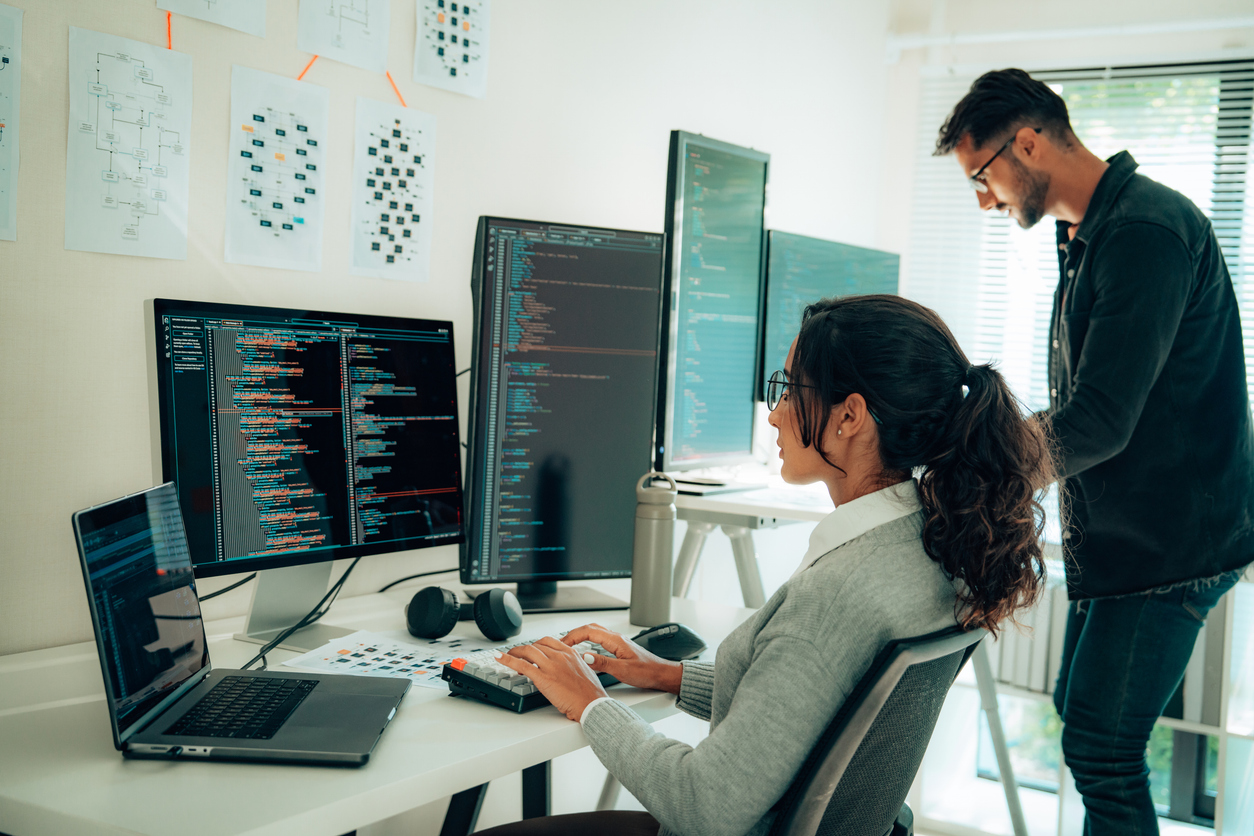 Two Cyber Professionals Reviewing Code on a Row of Monitors.