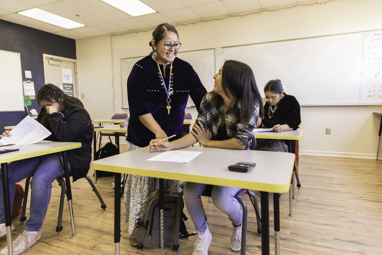 A Smiling Teacher Talks to a Teen Taking a Test Using a Calculator.