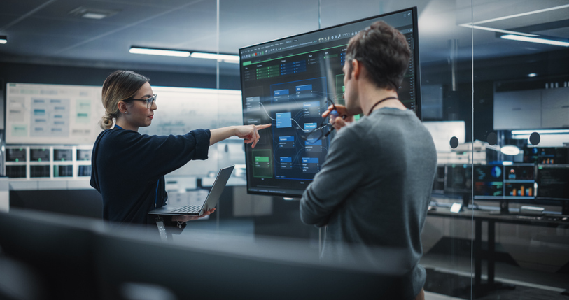 Network security engineer stands with a colleague next to a large monitor.