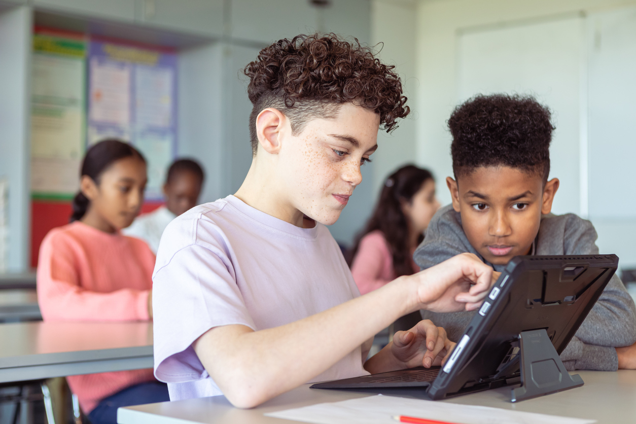 A Student Works on a Tablet in a Classroom While Another Student Watches.