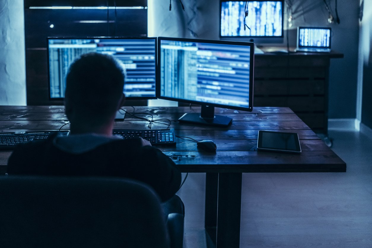 A Hacker Works in a Darkened Room Surrounded by Several Large Computer Monitors.