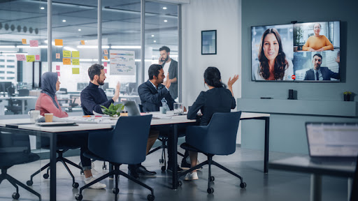 Members of a business team in an office meeting room collaborate with remote workers via video conference.