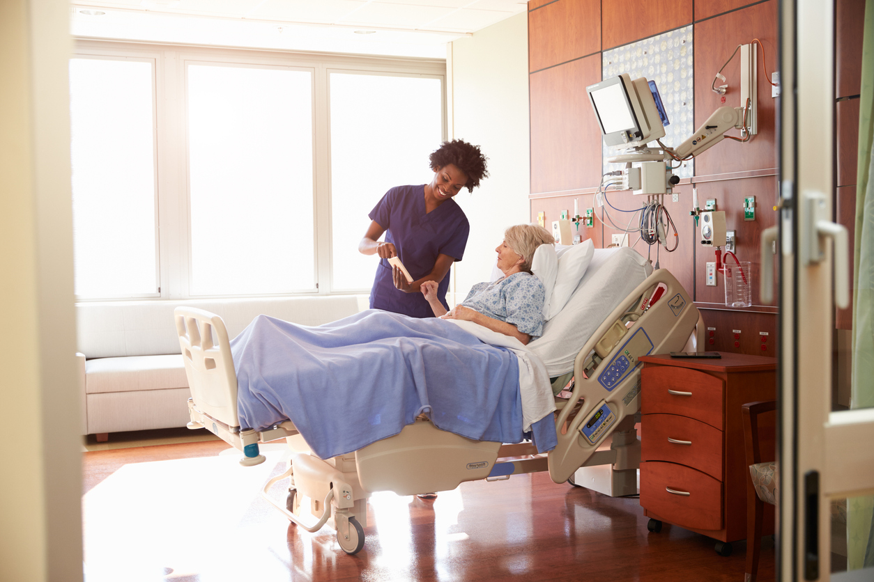 A nurse standing next to a patient’s hospital bed with a tablet. 
