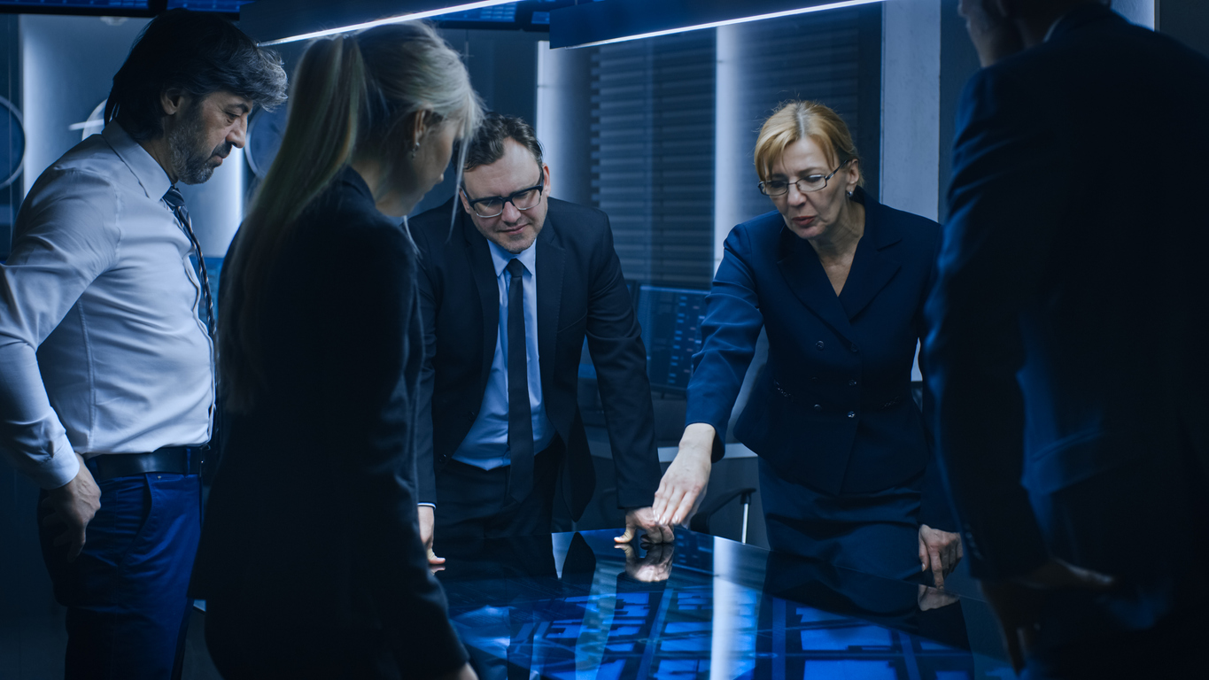 A team of federal agents meeting around an interactive digital table in a surveillance room.