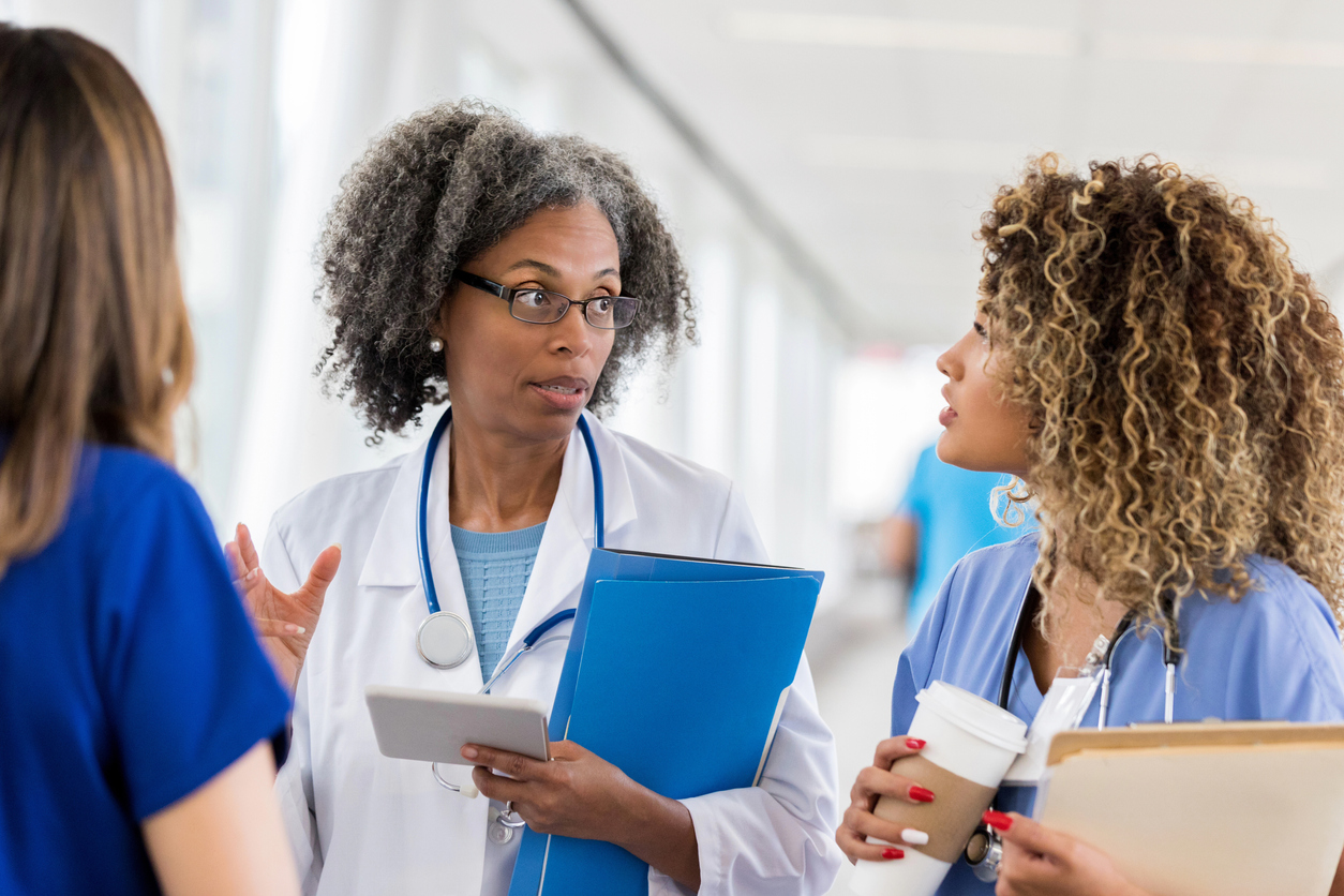 A Nurse Practitioner Speaks With Two Other Nurses in a Hospital Corridor.